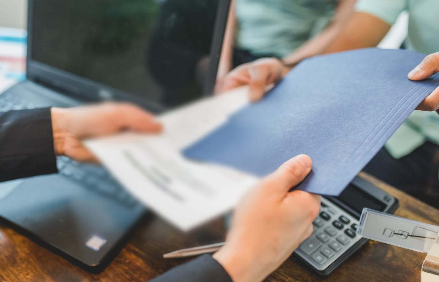 يدان تتبادلان ملفاً أزرق وأوراقاً فوق مكتب عليه حاسوب وآلة حاسبة
